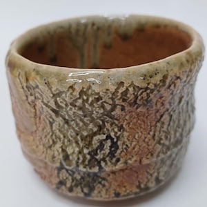 Puede incluir: Una taza de cerámica hecha a mano con un exterior texturizado. La taza presenta una superficie rugosa e irregular con una mezcla de tonos marrón, beige y verde oliva. El interior es de color marrón cálido y el borde tiene un acabado ligeramente brillante.