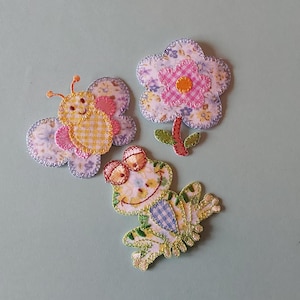 May include: Three colourful fabric appliques featuring a butterfly, a flower, and a frog. The butterfly is yellow and white with pink accents. The flower is pink with a yellow centre. The frog is green with blue and yellow accents.