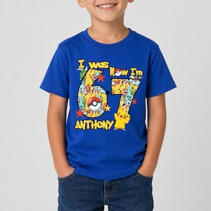 May include: A royal blue short-sleeve t-shirt featuring a graphic print of the number 67 filled with various cartoon characters and a Poké Ball. The text reads I was 6, Now I'm 7, and the name Anthony, accompanied by a yellow character.