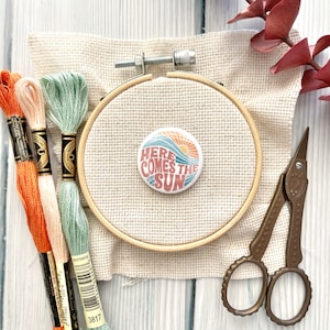 Puede incluir: Primer plano de un aro de bordado redondo con una tela beige estirada sobre él. Un botón con el texto "Here Comes The Sun" en una fuente retro está centrado en la tela. El botón tiene un gráfico de un sol y una ola. Tres carretes de hilo de bordar son visibles en la esquina inferior izquierda de la imagen.