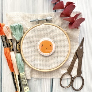 May include: A wooden embroidery hoop with beige fabric stretched across it. A smiling orange sun with white rays is centered on the fabric. Three spools of embroidery floss are on the left side of the hoop. A pair of bronze embroidery scissors are on the right side of the hoop.