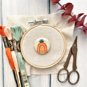 Könnte beinhalten: Nahaufnahme eines beigefarbenen Stickrahmens mit einem bunten Ananas-Design auf einem weißen Stoffhintergrund. Der Rahmen ist von Stickgarn und einer kleinen Schere umgeben.