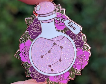 May include: A rose gold enamel pin featuring a glass bottle filled with pink glitter and the constellation Libra. The bottle has a white label that reads "Libra".