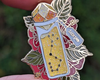 May include: Gold-tone enamel pin featuring a glass bottle filled with glitter and the constellation Scorpio. The bottle is topped with a multi-colored gem and surrounded by floral details.