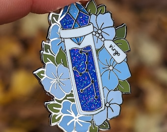 May include: A silver-colored enamel pin featuring a blue crystal vial with a constellation design, surrounded by blue flowers and green leaves. The tag reads "Virgo".
