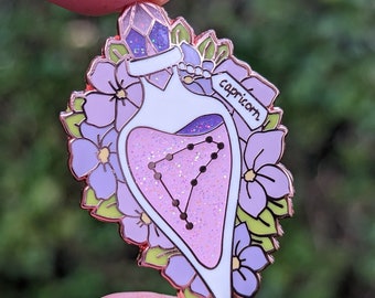 May include: A rose gold enamel pin featuring a heart-shaped bottle filled with pink glitter and the Capricorn constellation. The bottle is surrounded by purple flowers and green leaves. The tag on the bottle reads "Capricorn".