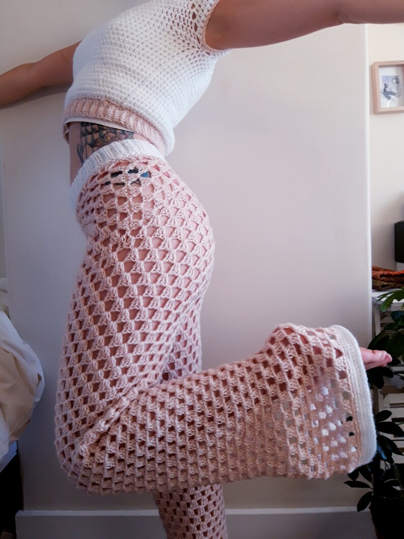 May include: A white crochet top and matching light pink crochet trousers. The trousers have a flared leg design. The top has a halter neck design. The crochet work has a diamond pattern.