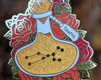 May include: Gold enamel pin featuring a glass bottle with a gold lid and a gold glitter fill. The bottle is surrounded by red roses and green leaves. The bottle has a white label with the word "Taurus" written in black.