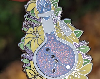 May include: A silver enamel pin featuring a blue glass bottle with a silver stopper and a silver tag that says "cancer". The bottle is filled with glitter and has a constellation inside. The bottle is surrounded by yellow, green, and purple flowers.