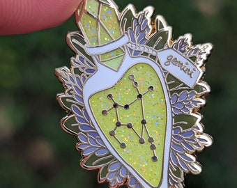 May include: Gold-tone enamel pin featuring a green and white heart-shaped bottle with a gold lid and a gold tag that says "gemini". The bottle contains a yellow glitter-like substance and a constellation design. The bottle is surrounded by green and purple leaves.