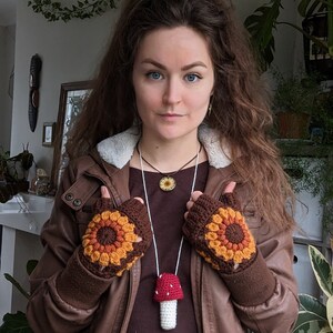 Peut inclure: Une femme portant une veste en cuir marron avec un gant sans doigts en crochet marron, orange et jaune sur chaque main. Elle porte un haut marron avec un collier en cordon blanc avec un pendentif en forme de champignon crochet&eacute;. Elle porte &eacute;galement une jupe blanche &agrave; motifs floraux.