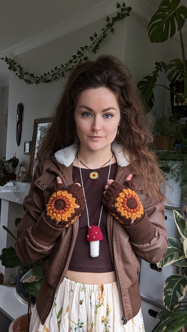 Peut inclure: Une femme portant une veste en cuir marron avec un gant sans doigts en crochet marron, orange et jaune sur chaque main. Elle porte un haut marron avec un collier en cordon blanc avec un pendentif en forme de champignon crochet&eacute;. Elle porte &eacute;galement une jupe blanche &agrave; motifs floraux.