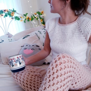 May include: A white crochet top and light pink crochet trousers. A floral-decorated mug is held near the lap. A gray cat-shaped pillow with a pink heart is also visible. The scene is set in a bedroom with floral decorations.
