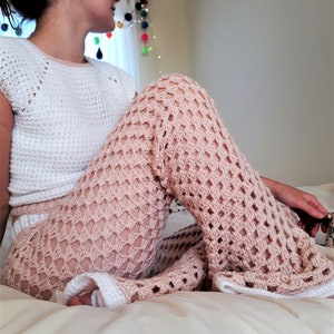 May include: A white crochet top with a scoop neck and short sleeves, worn with peach-coloured crochet trousers. The trousers have a flared leg design and an open diamond weave pattern. The handmade outfit is suitable for warmer weather.