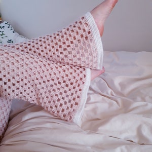 May include: Pale pink crochet sleeves with a square openwork design and white edging. The sleeves are on a bed with a cream-coloured duvet. The image has a soft, delicate feel.