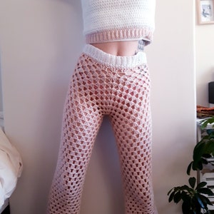 May include: A two-piece crochet outfit in white and pale pink. The top is a sleeveless crop top with a white body and a pale pink band. The trousers are a pale pink, open-weave crochet with a flared leg.