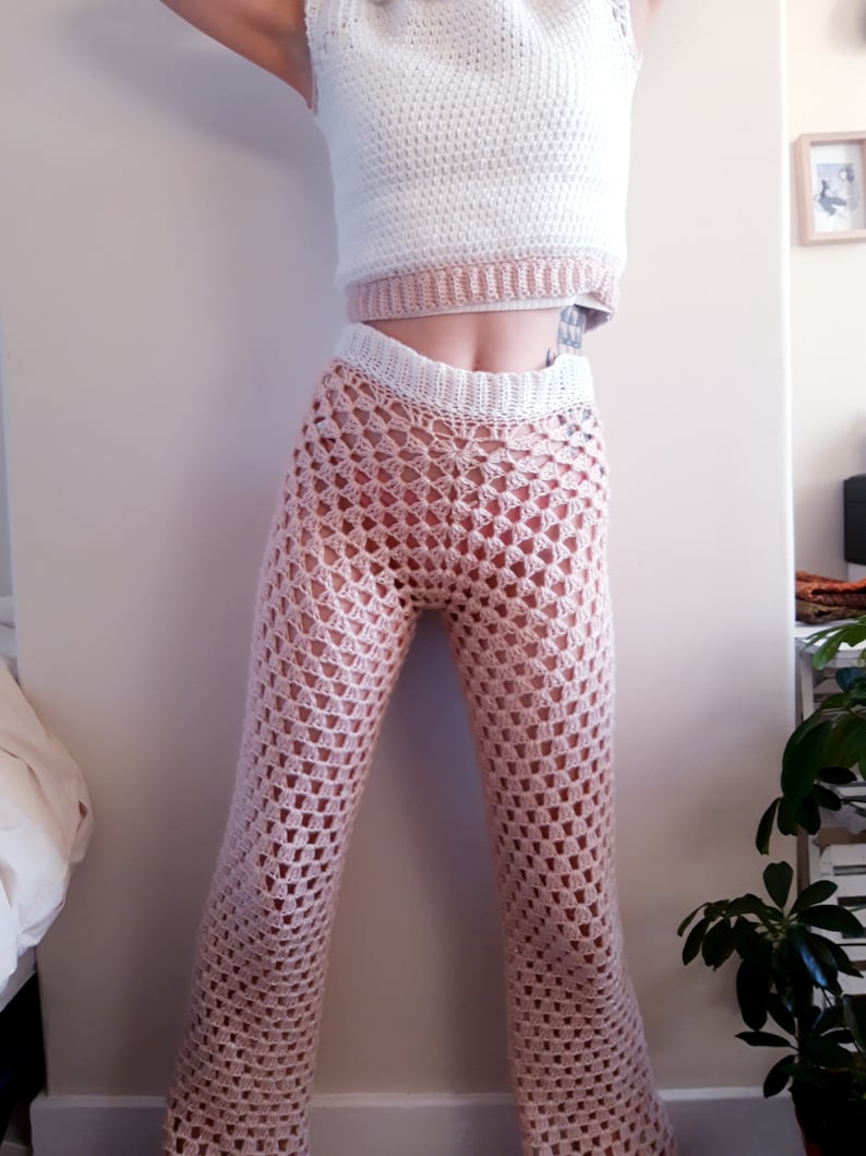 May include: A two-piece crochet outfit in white and pale pink. The top is a sleeveless crop top with a white body and a pale pink band. The trousers are a pale pink, open-weave crochet with a flared leg.