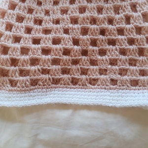 May include: Close-up of a peach-coloured crochet item with a grid-like pattern. The bottom edge features a white border. The handmade textile showcases a delicate, open weave design, perfect for summer wear.