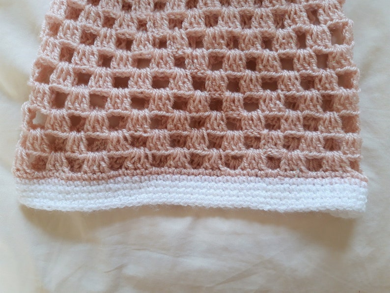 May include: Close-up of a peach-coloured crochet item with a grid-like pattern. The bottom edge features a white border. The handmade textile showcases a delicate, open weave design, perfect for summer wear.