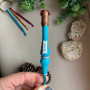 May include: A blue crochet hook with a brown dog figurine on top. The hook has a silver band with the word "Dog" on it and a white banner with the name "Arya" on it.