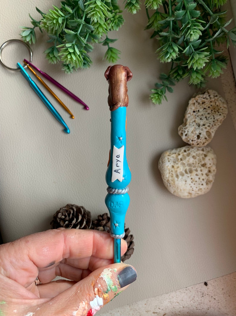 May include: A blue crochet hook with a brown dog figurine on top. The hook has a silver band with the word "Dog" on it and a white banner with the name "Arya" on it.