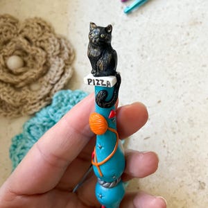 May include: A turquoise crochet hook with a black cat figurine sitting on a pizza box. The hook is decorated with a ball of yarn, a red heart, and a silver rope.