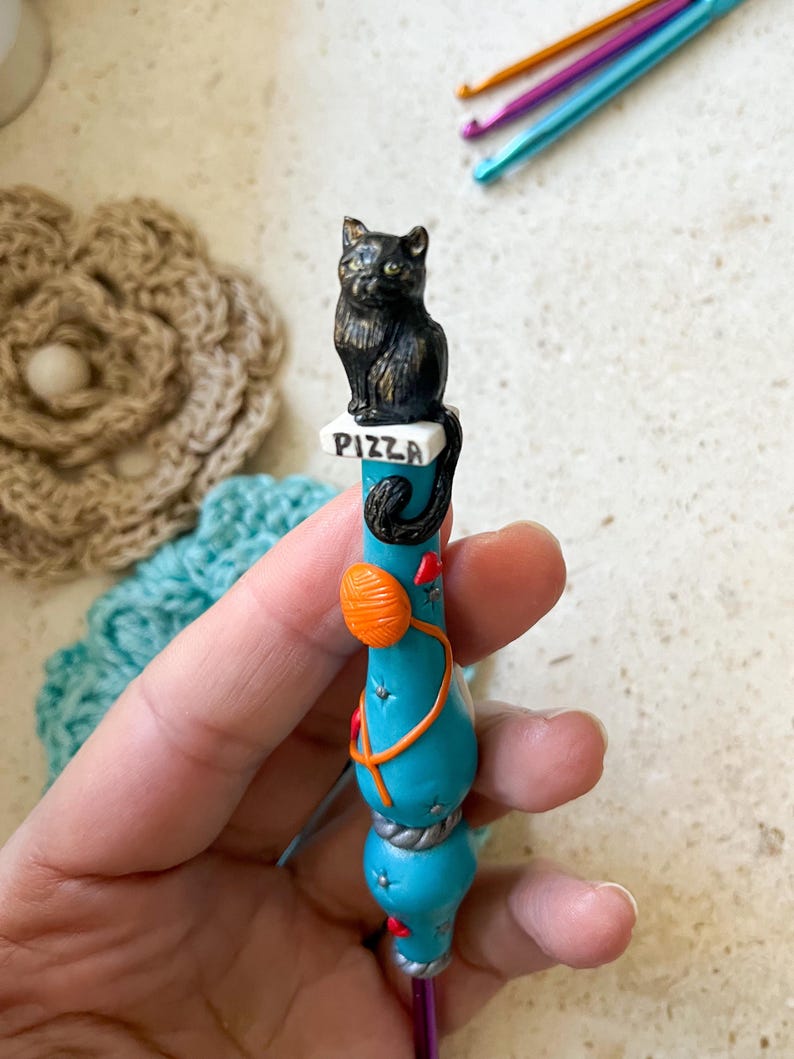 May include: A turquoise crochet hook with a black cat figurine sitting on a pizza box. The hook is decorated with a ball of yarn, a red heart, and a silver rope.