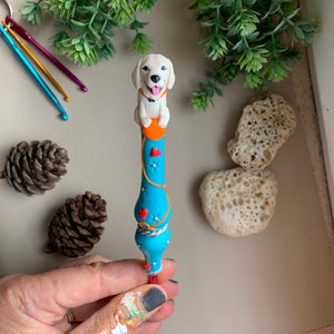 May include: A hand holds a blue and white crochet hook with a white dog figurine on top. The dog is wearing an orange collar and has a red heart on its chest. The crochet hook is decorated with red hearts and white stars.