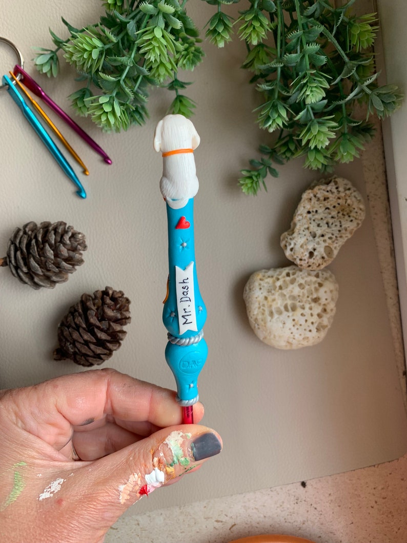 May include: A blue and white crochet hook with a white dog figurine on top. The hook has a label that says "Mr. Dash".