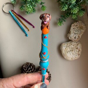 May include: A blue crochet hook with a brown dog figurine on top holding a ball of yarn. The hook is decorated with red hearts and orange yarn.