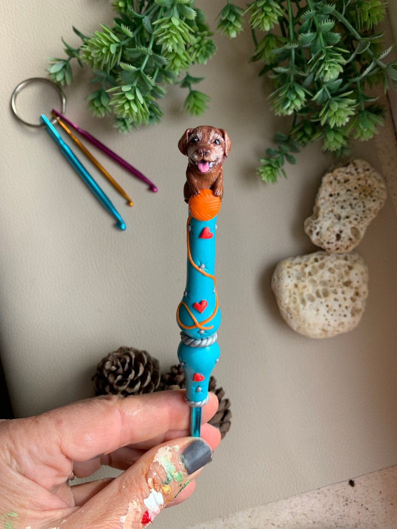 May include: A blue crochet hook with a brown dog figurine on top holding a ball of yarn. The hook is decorated with red hearts and orange yarn.