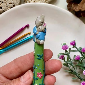 May include: A green crochet hook with a decorative handle featuring a bunny design. The handle is painted with vines, flowers, and bees. The bunnies are wearing blue jackets. Other crochet hooks are visible in the background.