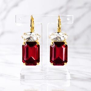 May include: A pair of gold-tone leverback earrings featuring a large, rectangular, deep red crystal stone topped with a smaller, clear, marquise-cut crystal. The earrings are suspended from a clear acrylic T-shaped display stand.