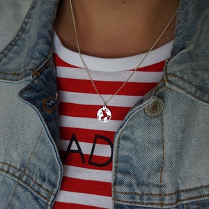 Puede incluir: Un collar de plata con un colgante de globo terráqueo. El collar lo lleva una persona que lleva una camisa a rayas rojas y blancas y una chaqueta vaquera.