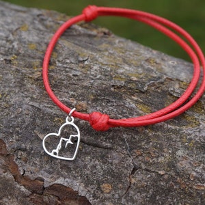 Puede incluir: Pulsera de cuerda roja con un dije de corazón plateado. El dije de corazón tiene un diseño recortado de una cordillera.