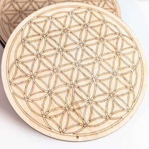 Könnte beinhalten: Zwei Holz-Untersetzer mit einem lasergravierten Lebensblume-Design. Die Untersetzer sind rund und haben eine hellbraune Farbe.