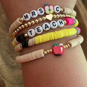 May include: Stack of beaded bracelets with teacher-themed charms. One bracelet spells out "MRS C" with an apple charm. Another says "TEACH" with a pencil charm. Other bracelets have gold beads and neutral-colored disc beads.