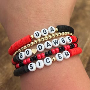 May include: A stack of beaded bracelets in red, black, and gold. The bracelets spell out "UGA", "DAWGS", "GO", and "SIC EM" with white letter beads. The bracelets are worn on a wrist.