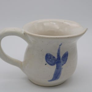 Puede incluir: Jarra de cerámica color crema con cuerpo redondeado y asa curva. Un diseño abstracto azul está pintado en la parte delantera. La jarra tiene un pico.