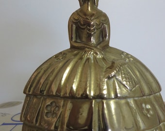 May include: A brass figurine of a woman in a dress, likely a trinket box. The figure has a hat and a detailed dress with floral accents. The brass has a polished finish, reflecting light.