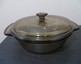 May include: A round, amber-colored glass casserole dish with a matching lid. The dish has two handles and a square knob on the lid. The glass has a slightly tinted appearance, and the dish appears to be vintage.
