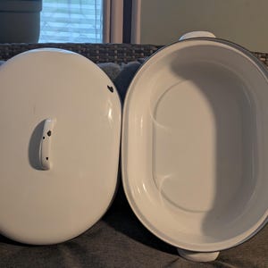 May include: An oval-shaped white enamel roasting pan with a matching lid. The lid features a central handle with small black decorative spots. The interior of the pan is smooth and white, with a dark grey rim around the top edge of the vessel.