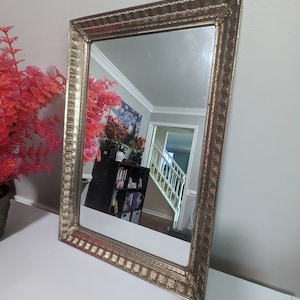 May include: A gold-toned rectangular mirror with an ornate frame. The frame has a repeating pattern of small squares and lines. The mirror is reflecting a room with a bookshelf and a staircase.