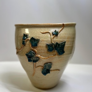 May include: A beige ceramic planter with a textured surface and a wide opening. The planter features raised teal-colored ivy leaves and vines. The design is a decorative accent for indoor or outdoor plants.