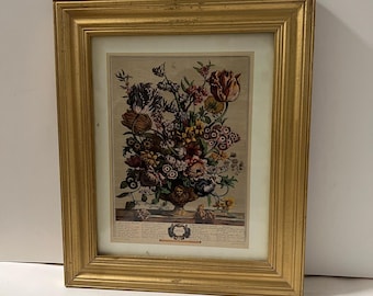 Puede incluir: Una impresión floral enmarcada que presenta una ilustración detallada de un ramo de flores en un jarrón decorativo. La impresión está enmarcada en un marco de madera dorada. La obra de arte incluye una variedad de flores en tonos rojos, rosas y amarillos.