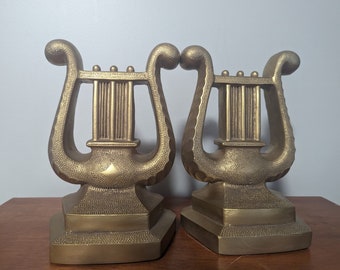 May include: A pair of gold-tone metal bookends shaped like lyres. The bookends have a detailed design and a textured surface.
