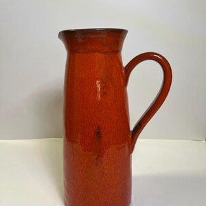 May include: A tall, glazed ceramic pitcher in a rich, reddish-orange hue. The pitcher features a rounded body, a curved handle, and a pouring spout. The interior rim is a darker brown. This handcrafted pitcher is suitable for serving beverages.