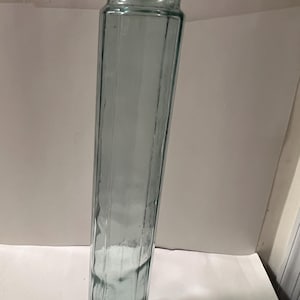 May include: A tall, clear, light blue glass vase with a rectangular shape and a round base. The vase has vertical ridges along its sides and a slightly flared top. The vase is empty and sits on a white surface.