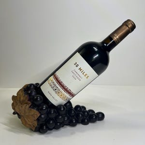 May include: A wine bottle rests in a decorative grape cluster wine holder. The bottle has a white label with the text "30 MILES Cabernet Sauvignon 2020 Napa Valley". The holder is black with brown leaves.