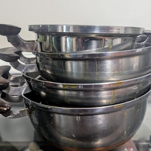 May include: A stack of stainless steel cooking pots with dark brown handles. The pots are of varying sizes and are stacked on top of each other. The handles are curved and appear to be made of wood or a similar material.